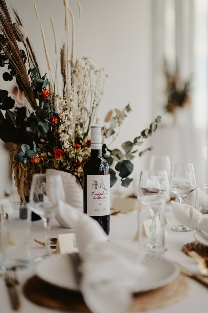 A table setting with a wine bottle, folded napkins, glasses, and a centerpiece featuring dried flowers and greenery.