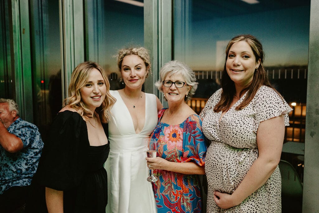 Four women pose together indoors, one holding a glass; a man is seated to the left. The room has large windows and city lights are visible outside at dusk.
