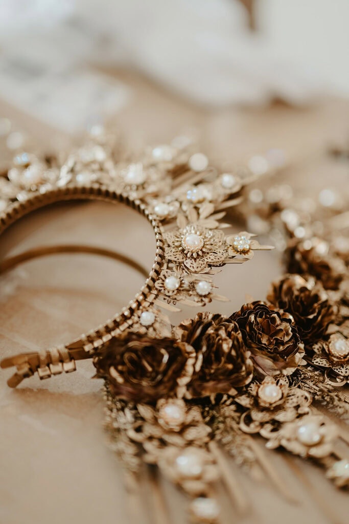 Close-up of an ornate golden headpiece adorned with intricate floral designs, pearls, and gemstones. The headpiece is placed on a light-colored surface.