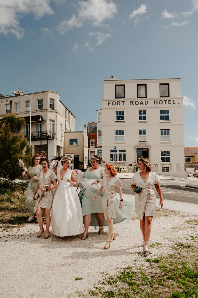 A bride and bridesmaids walk outside near the Fort Road Hotel on a sunny day, with some of the women laughing and chatting. The bride is in a white dress, and bridesmaids wear light-colored attire.