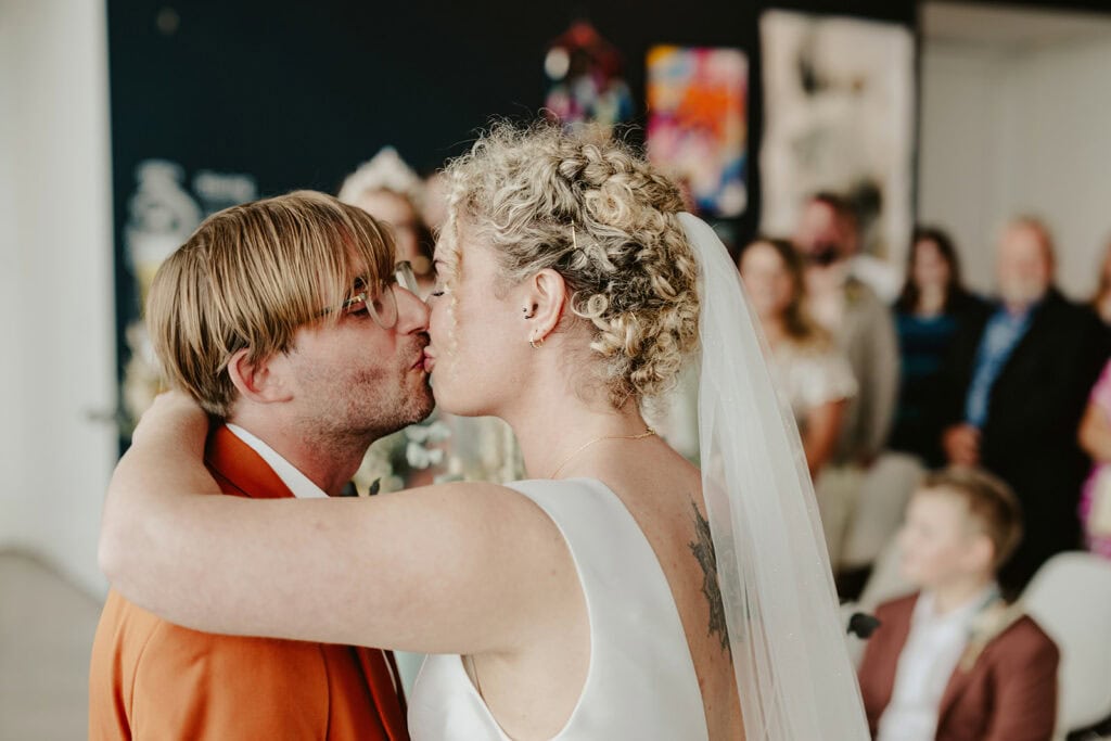 A couple shares a kiss during their wedding ceremony, surrounded by guests in a modern, art-filled venue.