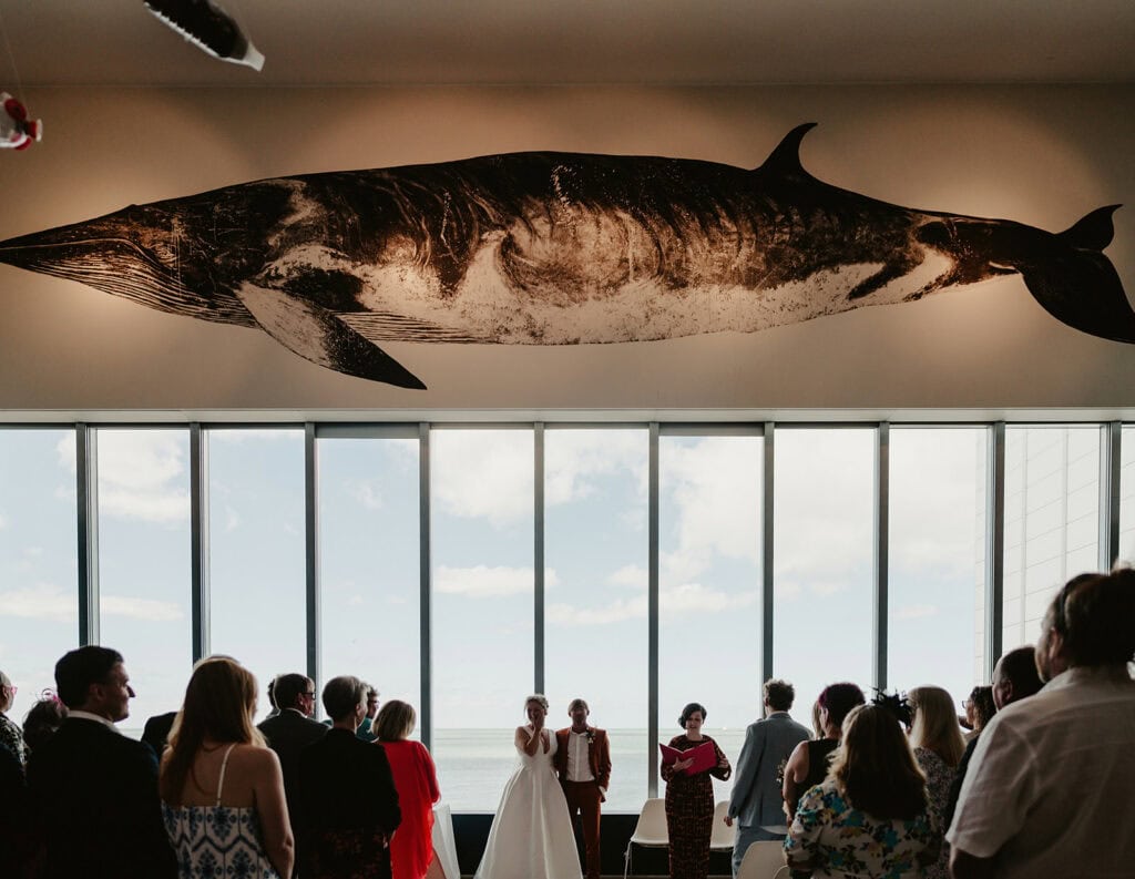 A wedding ceremony taking place indoors with a large whale sculpture mounted on the wall above the event and tall windows letting in natural light in the background.