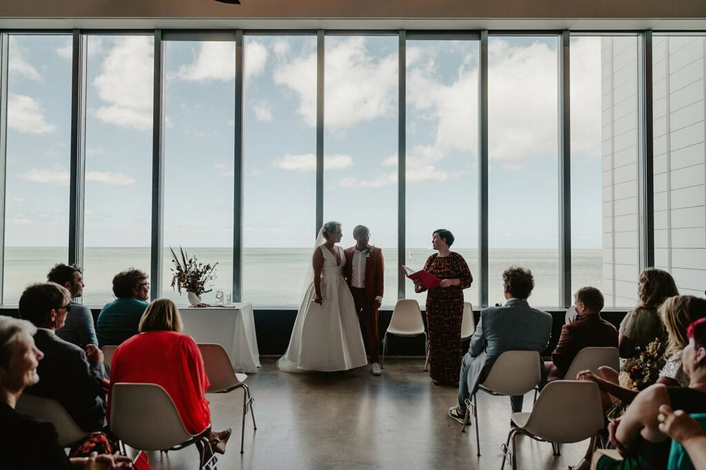 A couple stands in front of large windows overlooking the ocean at the Turner Contemporary, holding hands as an officiant reads from a book. Guests are seated on both sides, observing the ceremony.