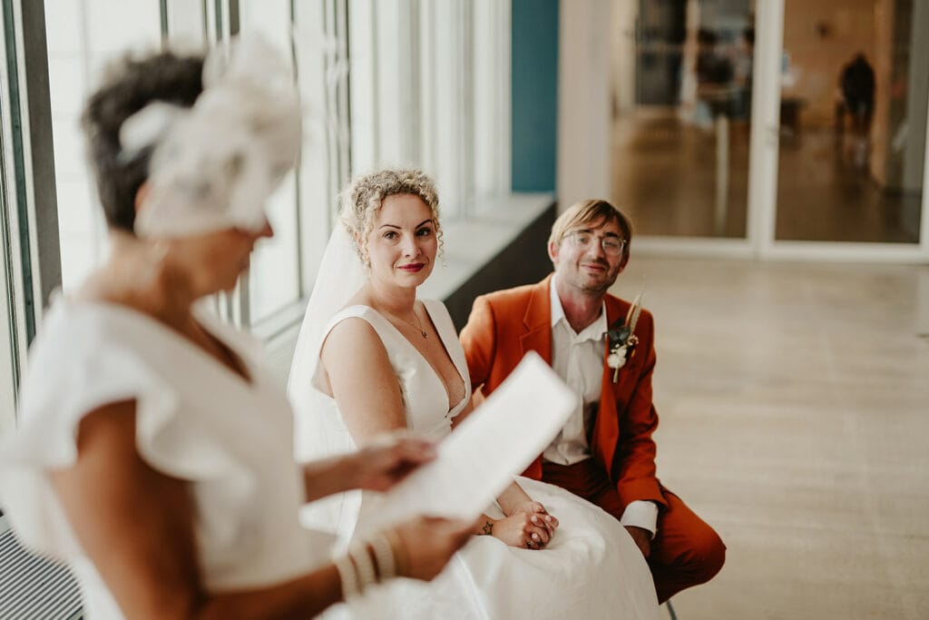 A bride and groom sit together, listening to a woman in a white outfit who is reading from a paper. The bride wears a white dress and veil, while the groom is dressed in an orange suit. They are indoors with large windows at Turner Contemporary in Margate, beautifully framing their wedding ceremony. Image by Pearce Wedding Photography.