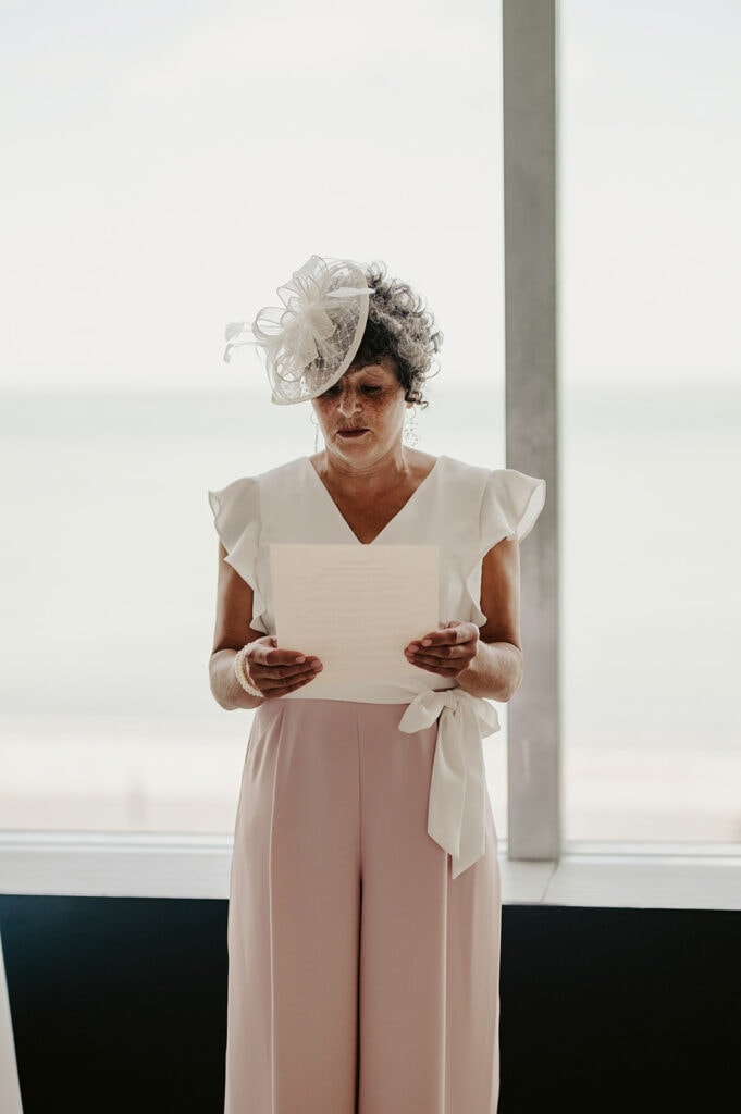 A person with a white fascinator reads from a piece of paper while standing in front of a large window with a beach view.