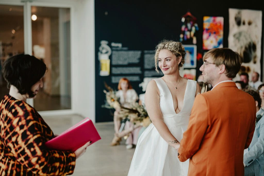 A couple stands facing each other, holding hands, during their wedding ceremony. A person officiates the ceremony, holding a pink folder. Colorful artwork is displayed on the walls in the background.
