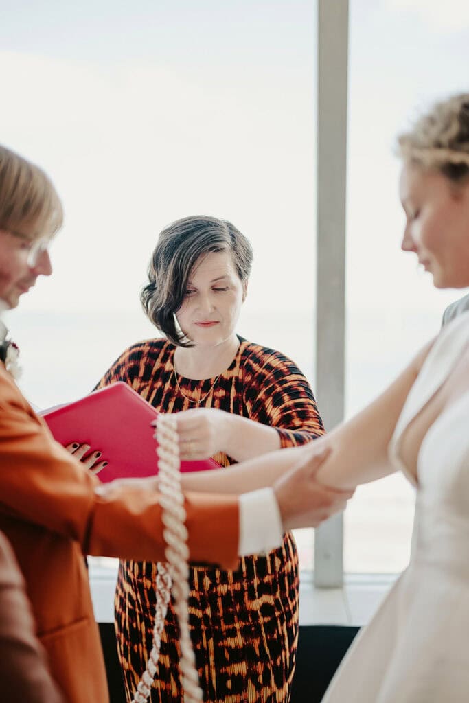 Three people participate in a handfasting ceremony; two of them are tying a knot around their wrists while the third person officiates, holding a red document.