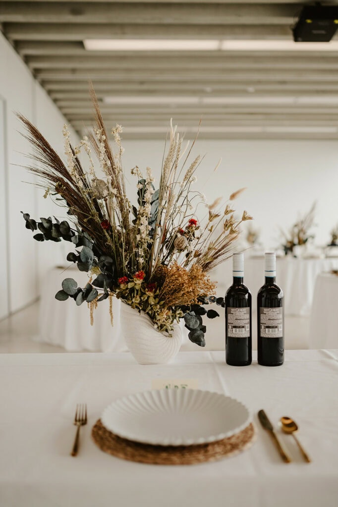 A dining table set with a large floral centerpiece featuring dried flowers and grasses in a white vase, reminiscent of the Turner Contemporary's artistic spirit. Two bottles of wine stand beside the centerpiece. The setting includes a white plate, gold utensils, and a wicker placemat, against a muted, contemporary background. Image by Pearce Wedding Photography.