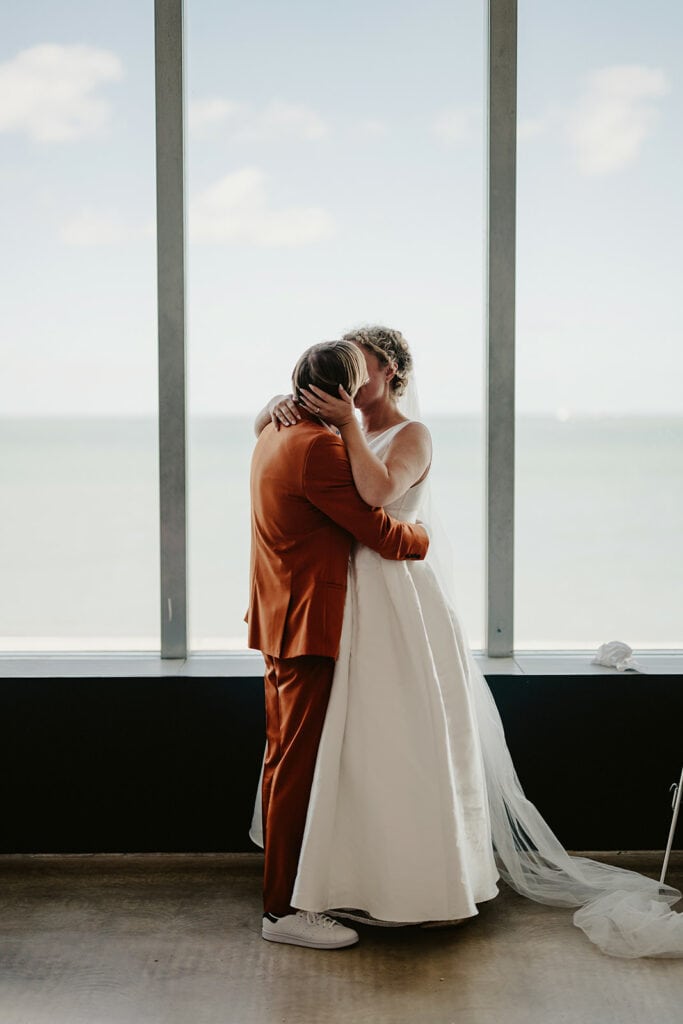 A couple embraces in front of a large window with an ocean view. The person on the left wears an orange suit, and the person on the right wears a white dress and veil.