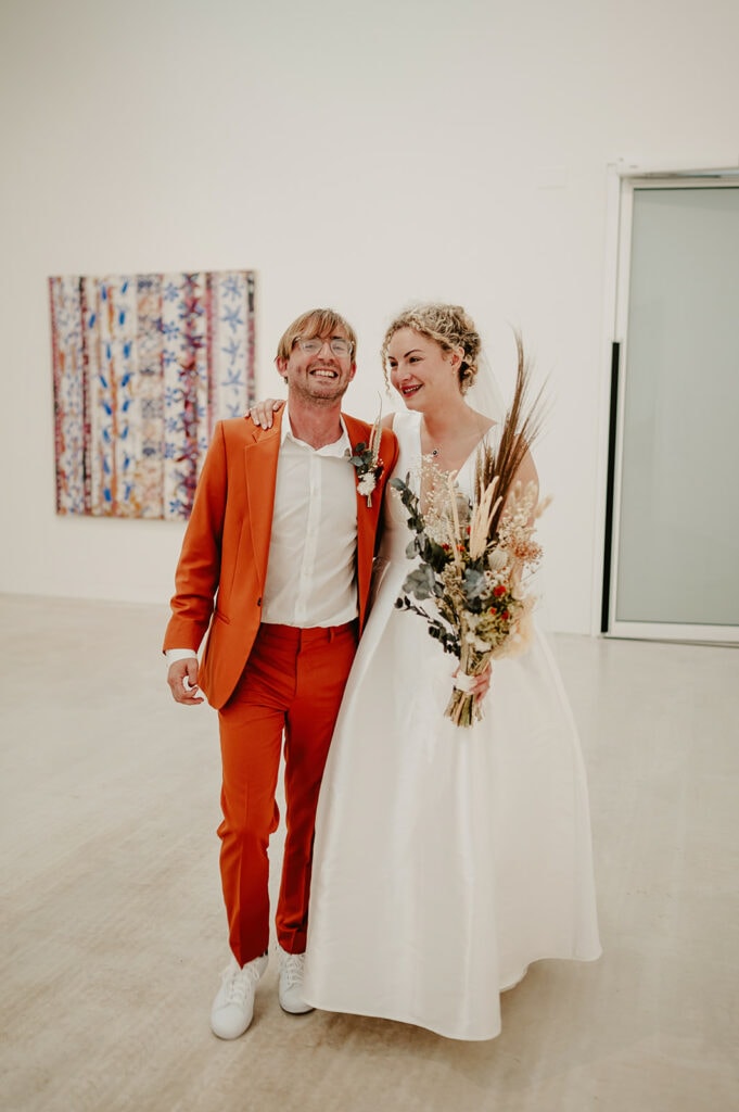 A smiling couple, dressed in an orange suit and white dress, stand close together in a minimalist room with modern art on the wall behind them. The woman holds a bouquet of mixed flowers and dried grasses.
