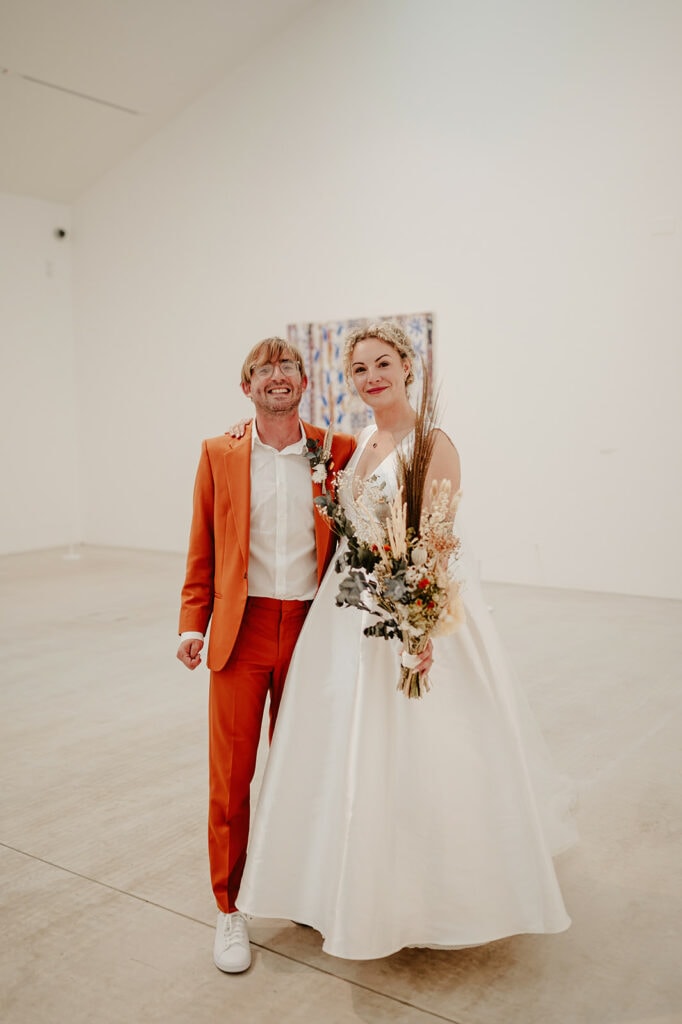 A couple, with the man in an orange suit and the woman in a white dress holding a bouquet, stands in a minimalistic gallery space with art on the wall behind them.