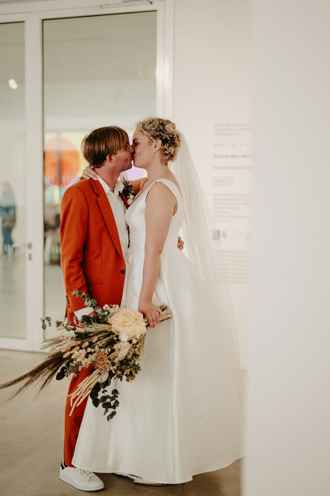 A couple dressed in wedding attire, with the groom in an orange suit and the bride in a white gown, kiss while holding a bouquet of flowers in front of a glass door.