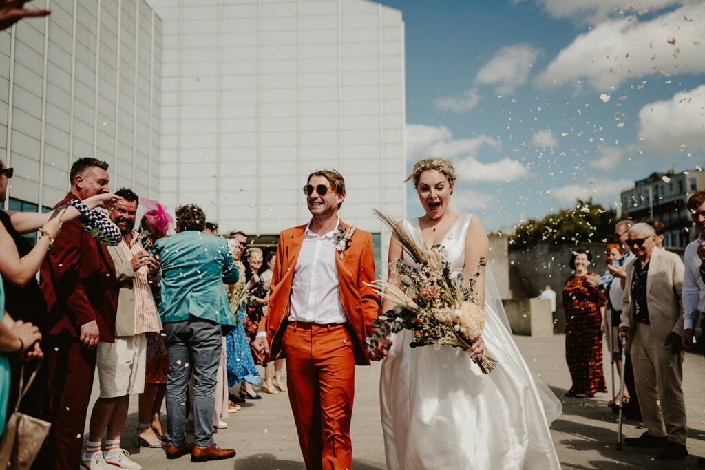A newlywed couple joyfully walks hand in hand through a celebratory crowd under a sunny sky at Turner Contemporary in Margate. The groom is in an orange suit and sunglasses, while the bride wears a flowing white dress, holding a bouquet. Guests shower them with confetti as they smile and cheer. Image by Pearce Wedding Photography.