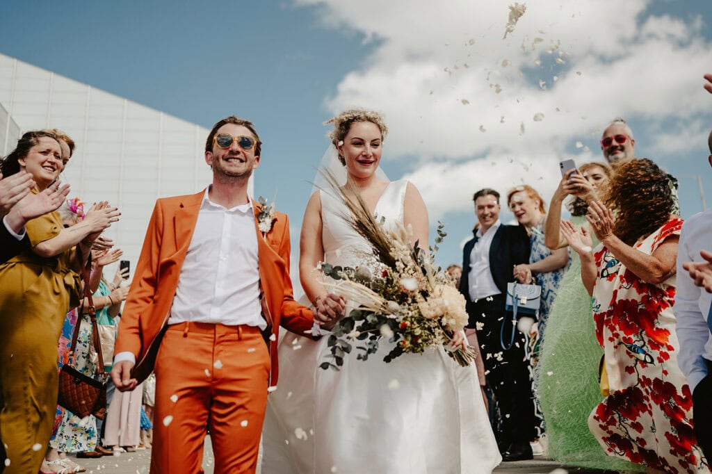 A bridal couple walks outside, hand in hand, as guests surround them throwing confetti. The groom wears an orange suit, and the bride wears a white gown, both smiling amid the celebration.