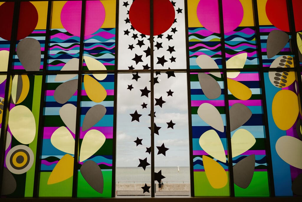 A large, colorful stained glass window at Turner Contemporary in Margate features abstract shapes in vibrant colors such as pink, purple, blue, and yellow. The center panel highlights a bold red circle above a series of black stars. Behind the window, a scenic beach and ocean can be seen—perfect for wedding photos. Image by Pearce Wedding Photography.