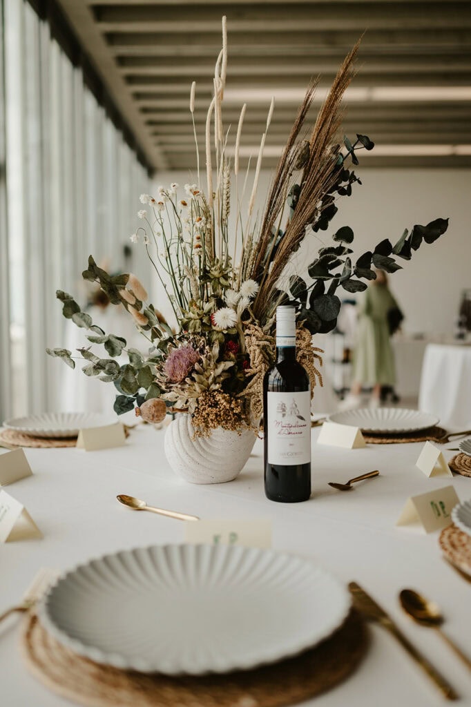 A table set for a formal occasion, perhaps a wedding, features a centerpiece with a wine bottle and a floral arrangement of various dried plants in a white vase. The table is adorned with white plates, woven placemats, and gold-colored cutlery against the backdrop of large windows reminiscent of Turner Contemporary in Margate. Image by Pearce Wedding Photography.