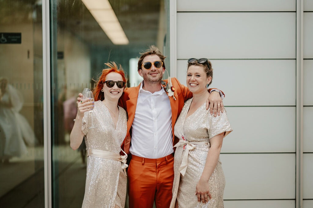 Three people are standing together, smiling at the camera. The person in the middle is wearing an orange suit, while the two on either side are wearing sparkly, light-colored dresses.