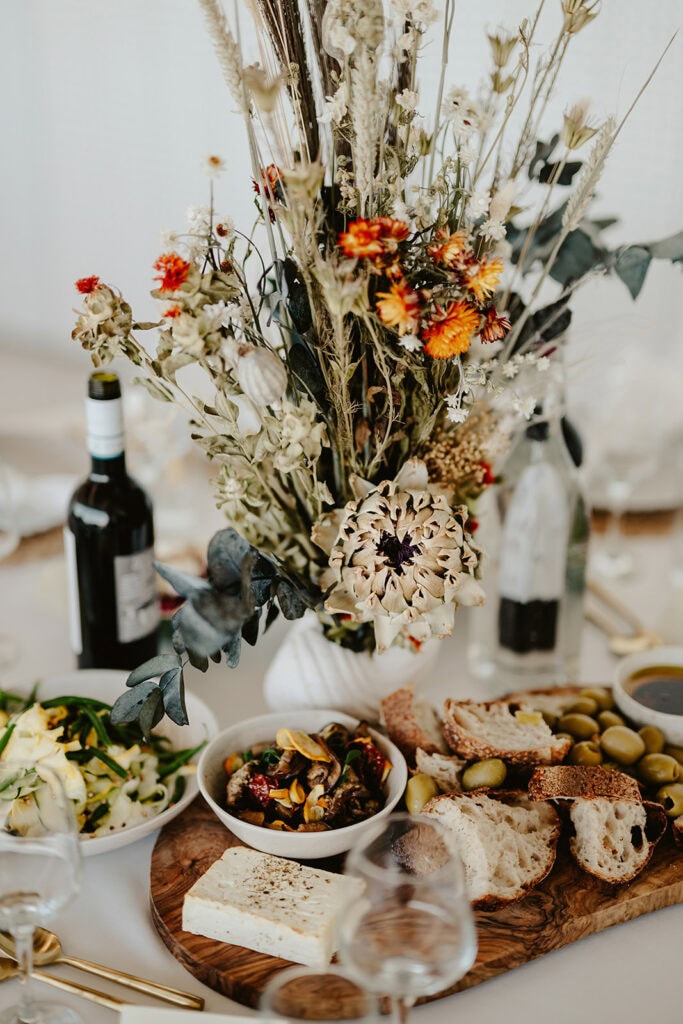A table set with bread, cheese, olives, a salad, and two bottles (one possibly wine), with a centerpiece of mixed flowers.