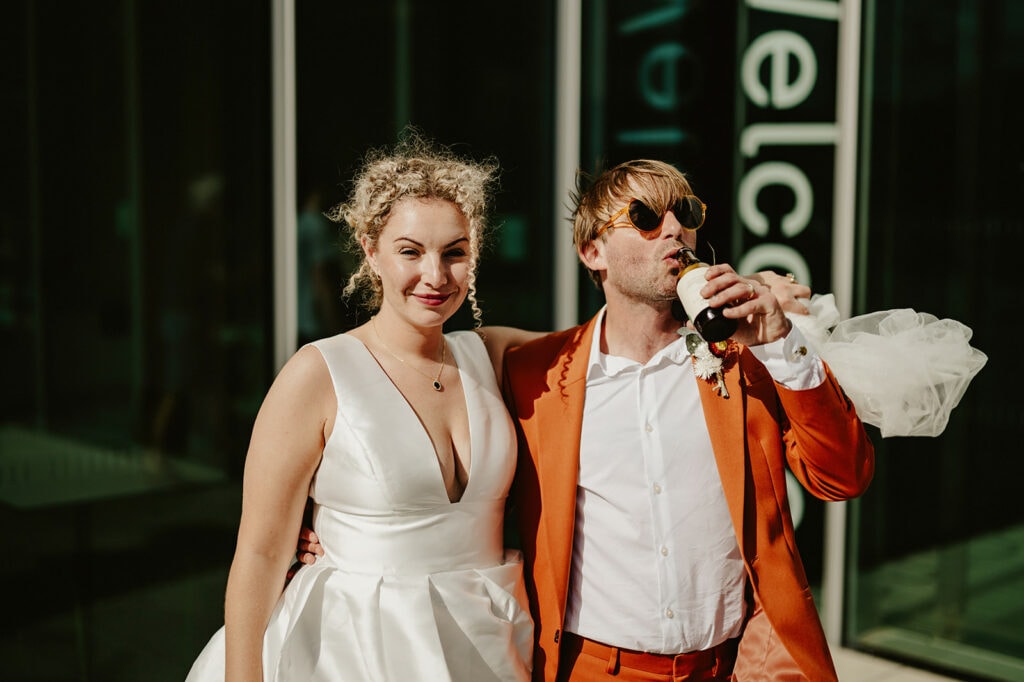 A bride in a white dress and a groom in an orange suit stand together outdoors. The groom is drinking from a bottle while the bride smiles at the camera.
