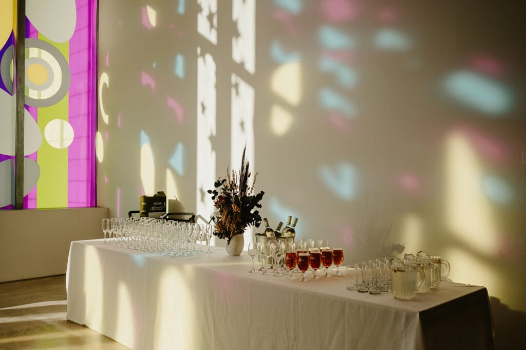 A table set with glasses of champagne and wine, pitchers, and floral centerpieces in a room with colorful stained glass windows casting abstract light patterns on the walls.
