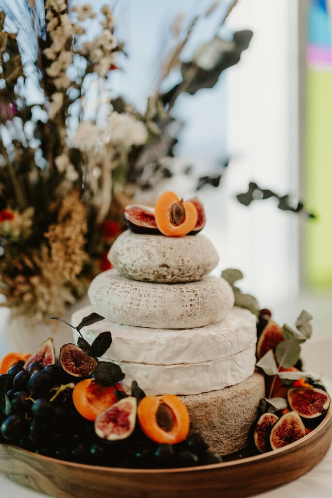 A tiered cheese cake garnished with halved apricots, figs, and green leaves is displayed. A bouquet of dried flowers is in the background.