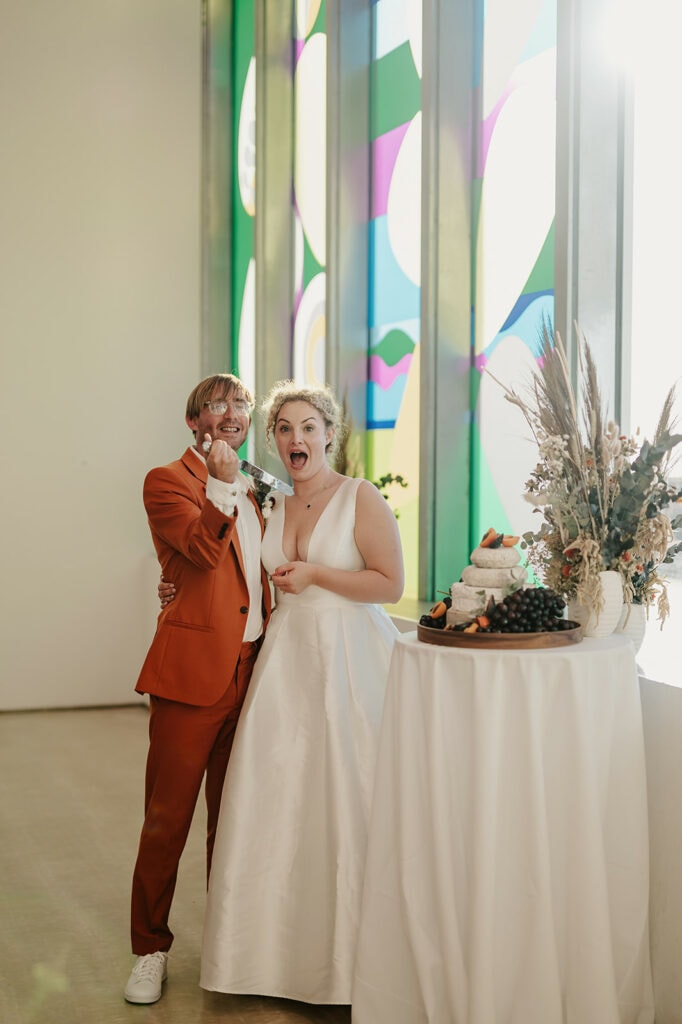 A couple stands by a table with a wedding cake. The woman in a white dress and the man in an orange suit are smiling and looking at the camera, holding their left hands up with the rings visible.