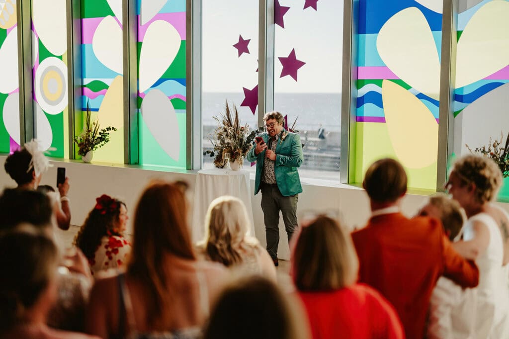 A person wearing a green jacket and holding a microphone addresses a crowd at an indoor event at Turner Contemporary in Margate. The venue features large windows with colorful star and abstract pattern decorations, and sunlight streams through, creating a vibrant atmosphere. Image by Pearce Wedding Photography.