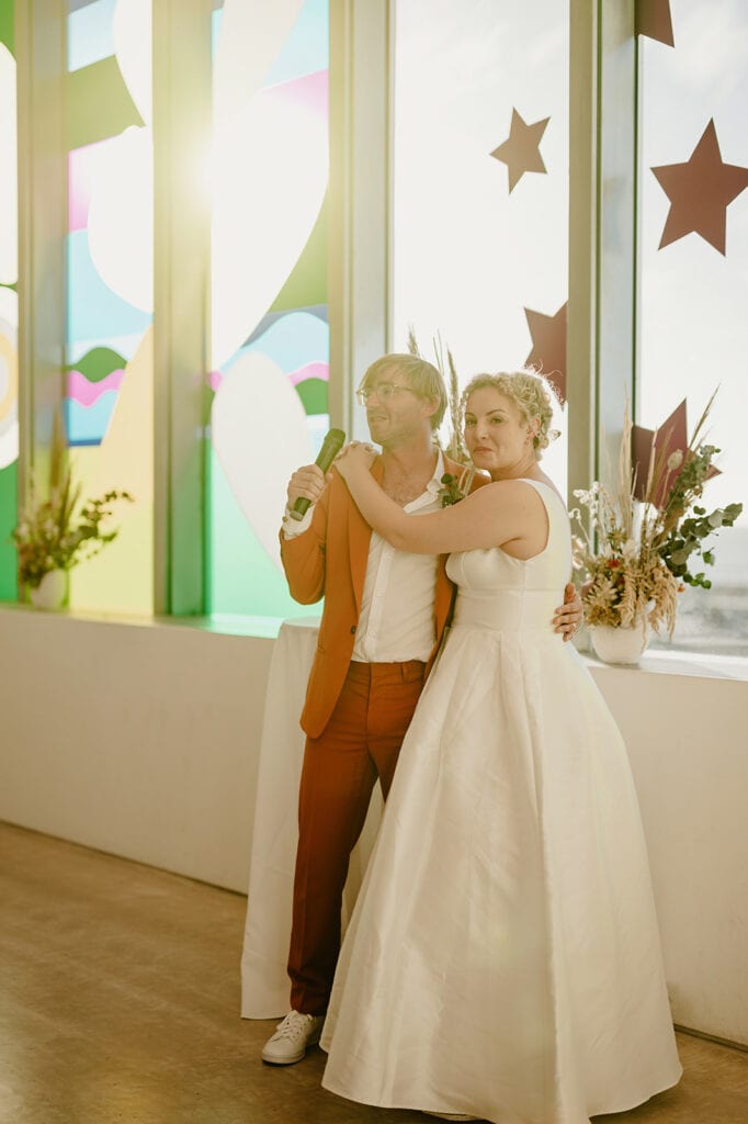 A couple stands together in wedding attire, the man holding a microphone. They are in front of large windows decorated with colorful designs and star-shaped cutouts.