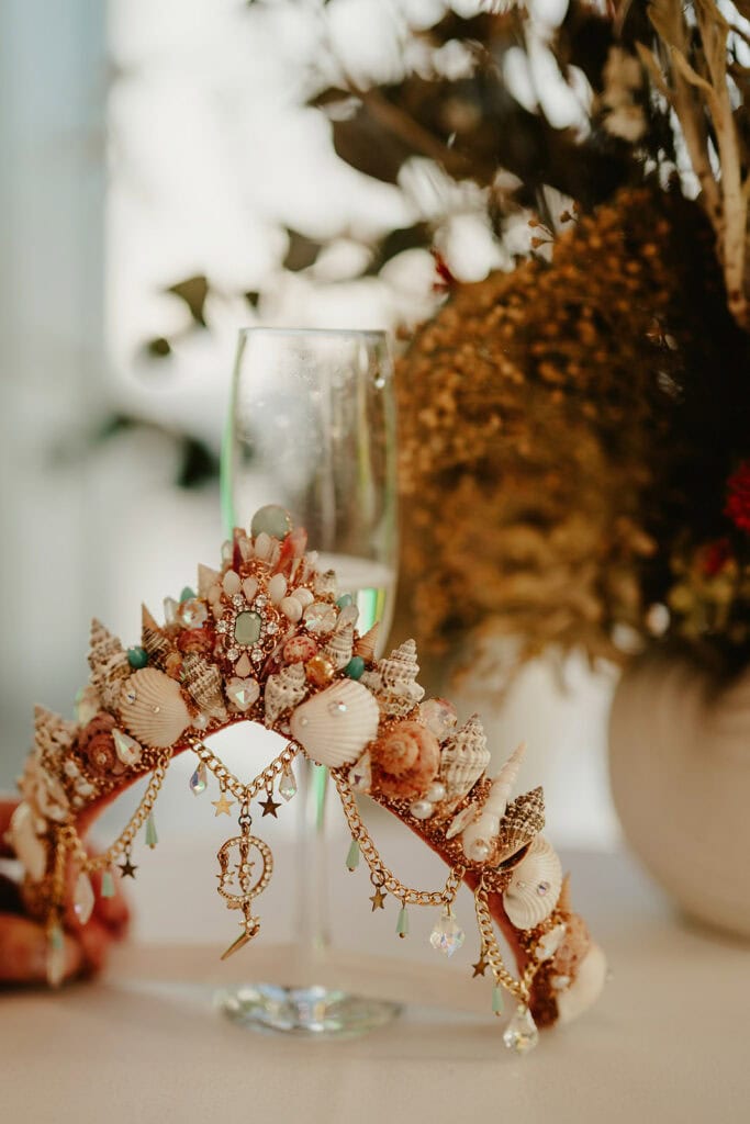 A decorative headpiece adorned with seashells, crystals, and dangling charms rests on a table in front of a champagne flute and a vase with dried flowers.