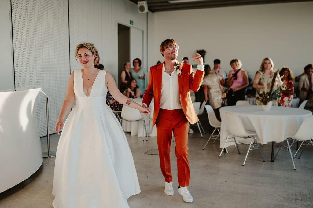A smiling couple in wedding attire walks hand-in-hand; the bride is in a white dress and the groom is in an orange suit. Guests are seated and standing in the background.