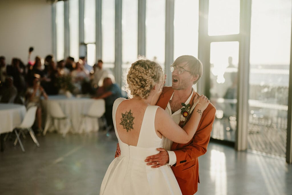 A couple dances in a sunlit room with large windows, while guests in the background watch. The woman has a tattoo on her back, and the man wears glasses and an orange jacket.