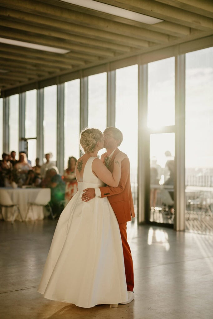 Two people dance closely together in a sunlit room with large windows. One person wears a white dress, and the other wears an orange suit. Guests, seated at tables in the background, watch the couple. The setting appears to be a wedding celebration at Turner Contemporary in Margate. Image by Pearce Wedding Photography.