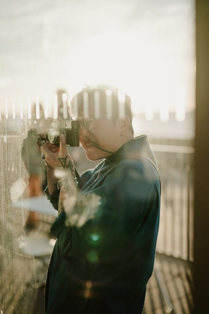 Person with short hair and tattoos wearing glasses and a blue jacket, taking a photograph through a glass wall in a sunlit outdoor setting.
