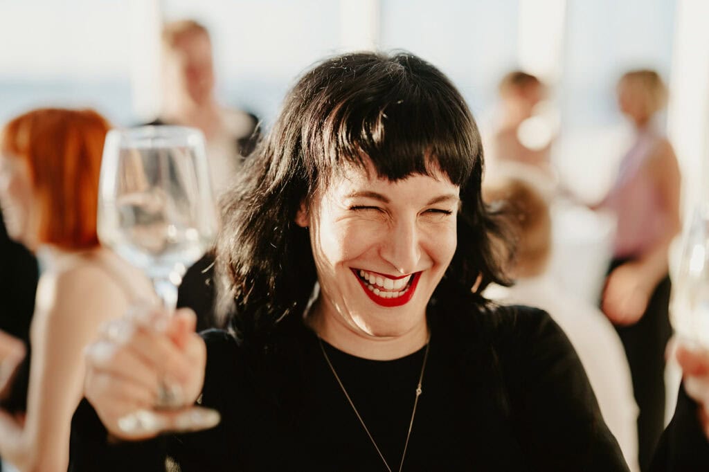 A woman with black hair and red lipstick smiles widely while holding two wine glasses at a social event.