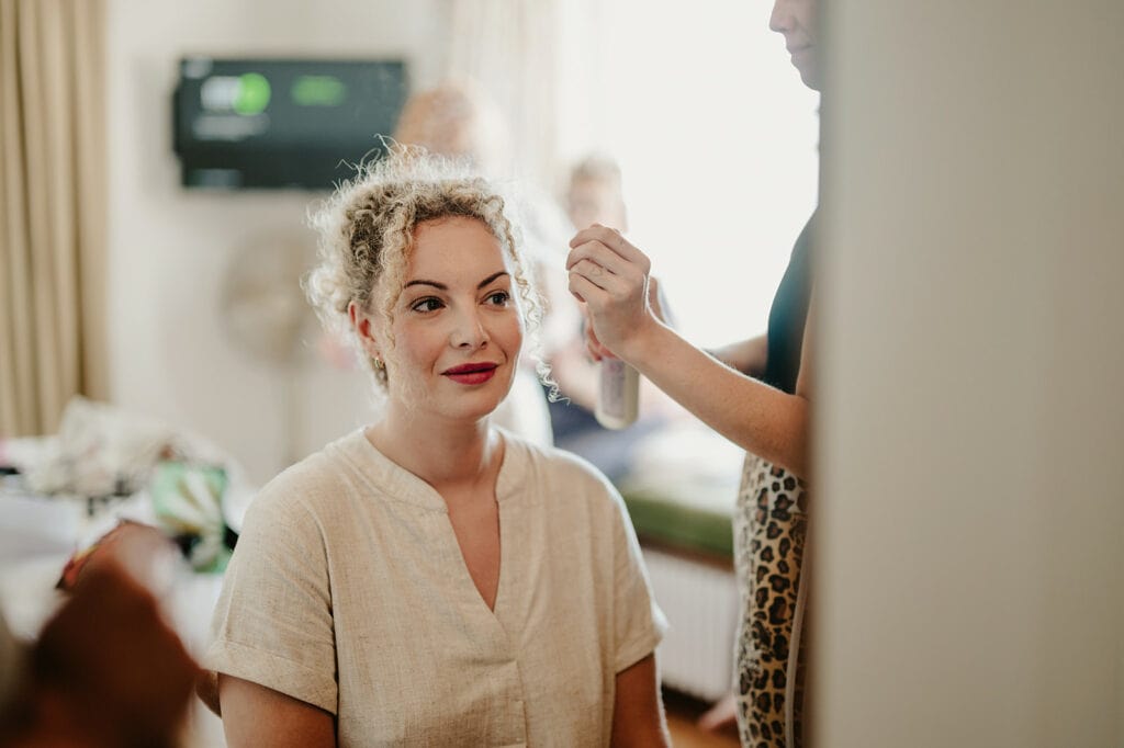 A woman with curly hair sits while another person styles her hair, holding a spray bottle. She is wearing a light-colored shirt, and the setting appears to be a room with various items in the background.