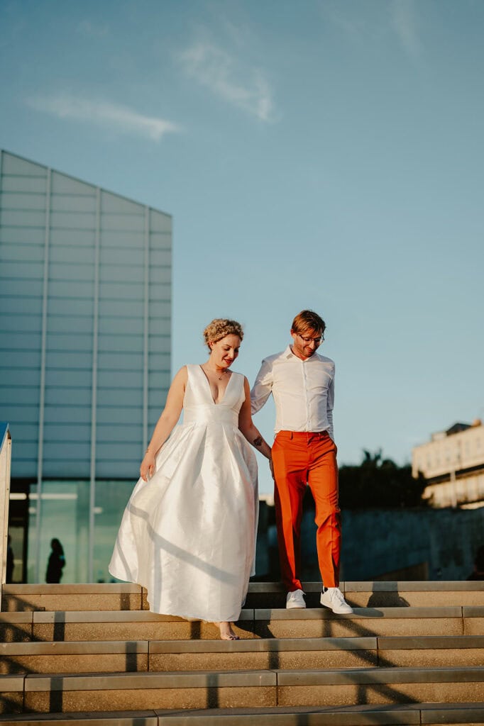A couple, with the woman in a white dress and the man in a white shirt and orange pants, walk down a set of stairs in an urban area with modern architecture in the background.