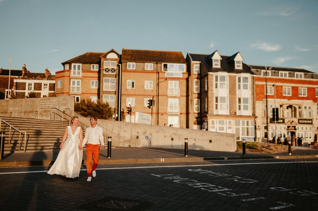 A couple in wedding attire walks hand in hand near a seaside town with red-brick buildings in the background during sunset.