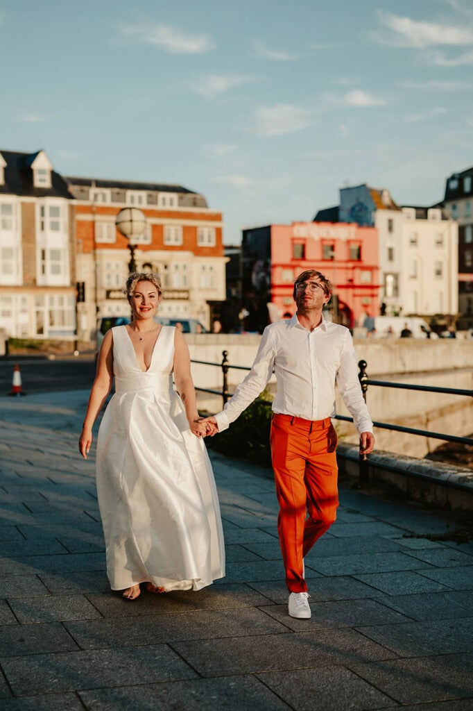 A couple dressed in formal attire—white dress and orange pants—walks hand in hand on a city street with buildings and a blue sky in the background.