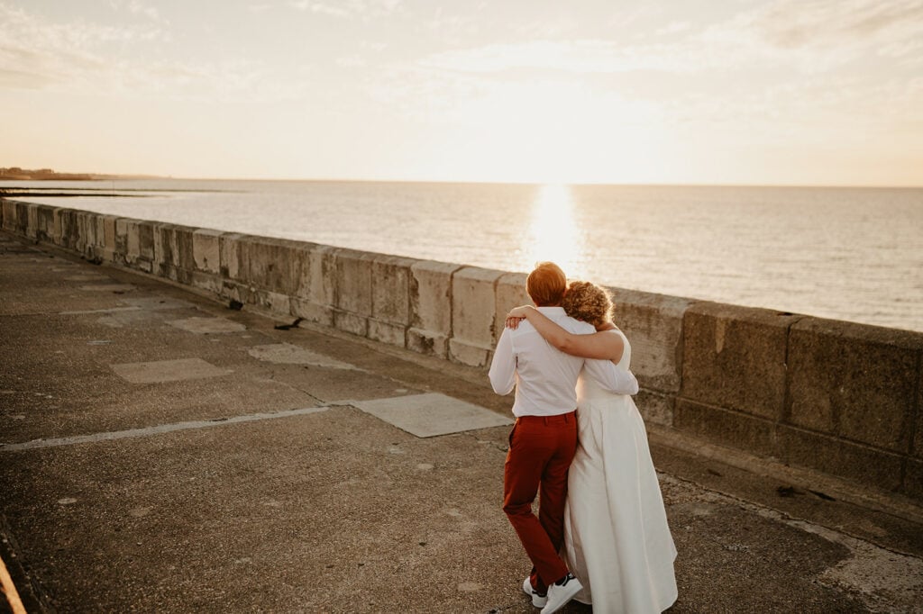 Two individuals embracing while walking along a seaside promenade at sunset, with the sun setting over the horizon and casting a warm glow on the scene.