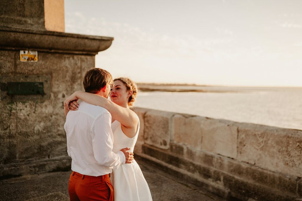 A couple embraces outdoors near a stone wall at sunset, with a scenic view of water in the background.