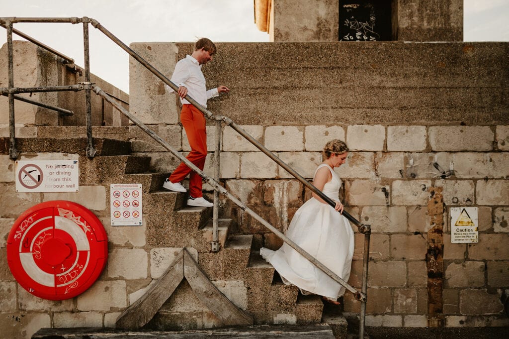 A man and woman dressed in formal attire descend a worn stone staircase with a lifebuoy and caution signs on the wall beside them.