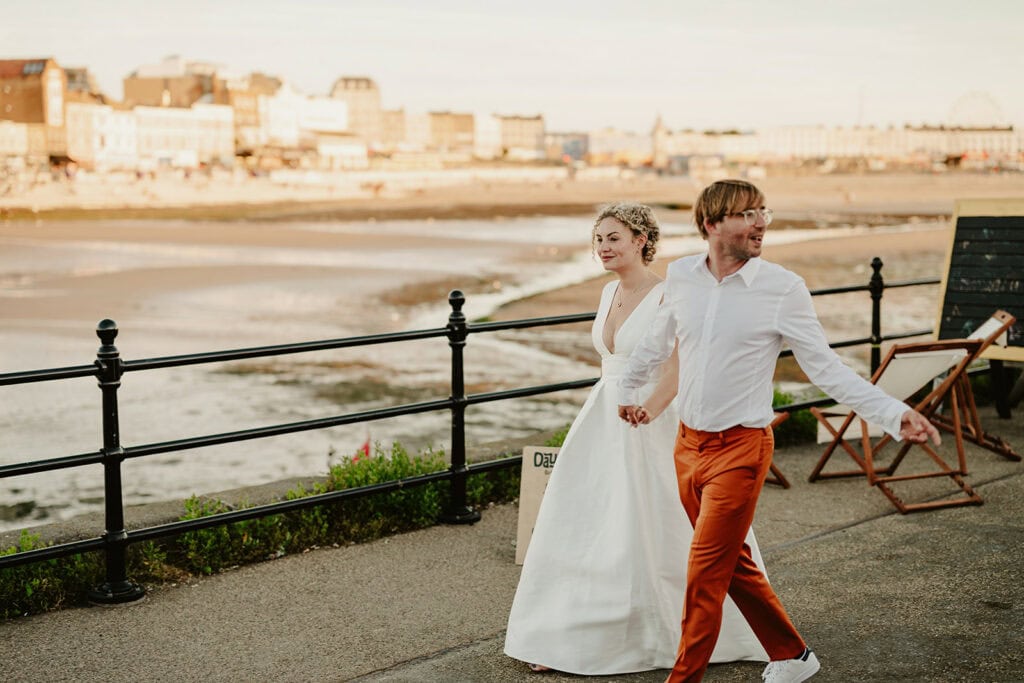 A couple, dressed in wedding attire, holds hands as they walk along a seaside promenade with a town and beach in the background.