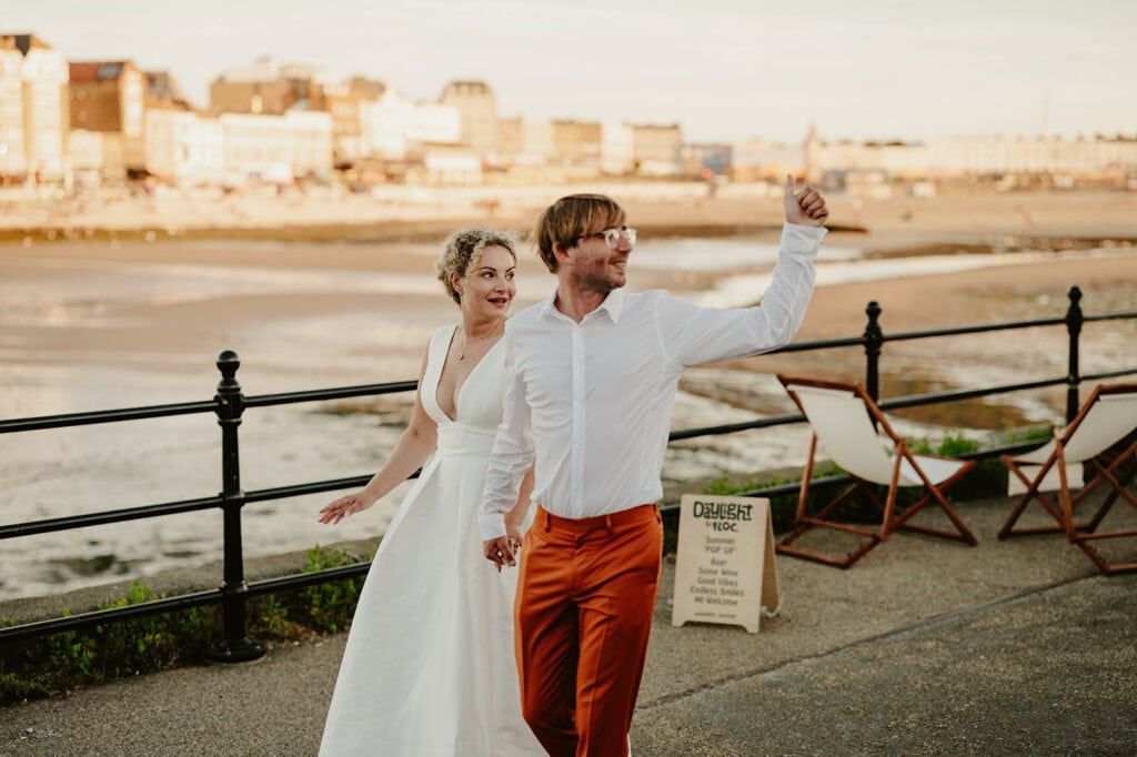 A couple dressed in formal attire, with the woman in a white dress and the man in an orange suit, walk hand in hand on a beachside promenade. The man is gesturing excitedly with his hand.
