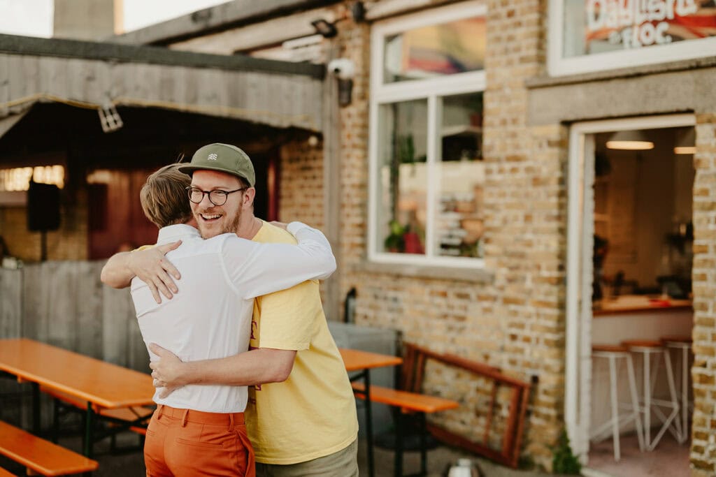 Two individuals embrace each other joyfully outside a brick building with a partially visible sign. The area has orange picnic tables and stools inside. One person wears an orange outfit, the other a yellow shirt and cap.