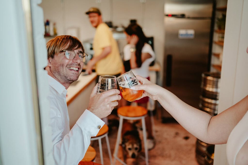 Two people clink beer glasses in a kitchen, with two others preparing food in the background. A dog sits on the floor.