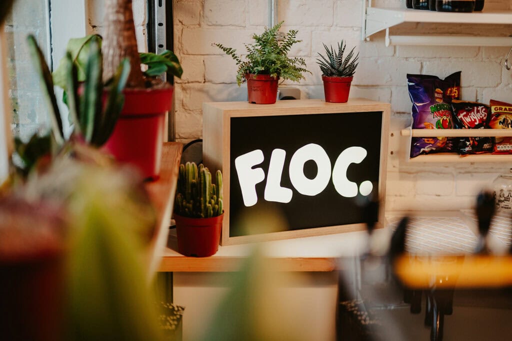 A brightly lit sign reading "FLOC." sits on a shelf surrounded by plants and snacks in a cozy, white-brick room.