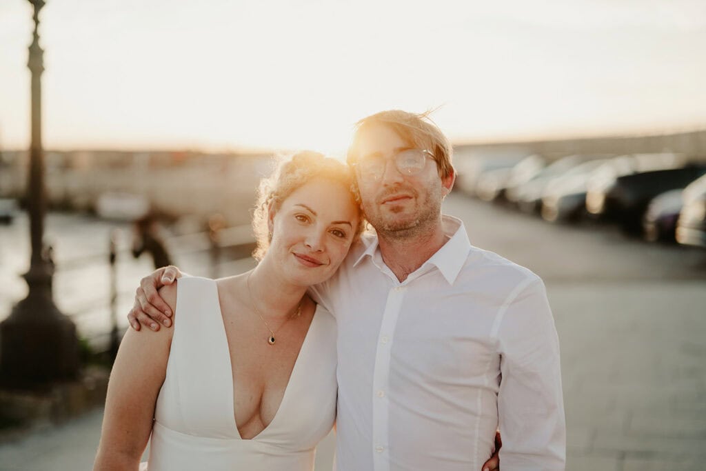 A couple stands outdoors at sunset with their arms around each other, dressed in white; the woman in a sleeveless dress and the man in a button-up shirt. Smiling warmly amid parked cars and a waterfront, they resemble newlyweds celebrating near Turner Contemporary, adding Margate's charm to their moment. Image by Pearce Wedding Photography.