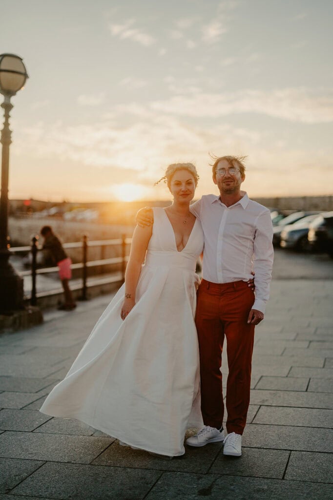 A couple stands arm in arm at sunset, the woman in a white dress, and the man in a white shirt and red pants. They are on a paved walkway with a lamp post and railing in the background.