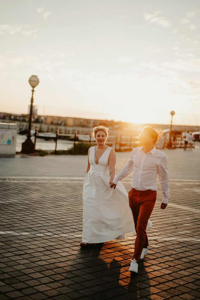 A couple dressed in wedding attire walks hand in hand along Margate's waterfront promenade at sunset. The bride wears a white dress, and the groom is in a white shirt and brown pants. The sun casts a warm glow near the Turner Contemporary, creating a romantic atmosphere. Image by Pearce Wedding Photography.