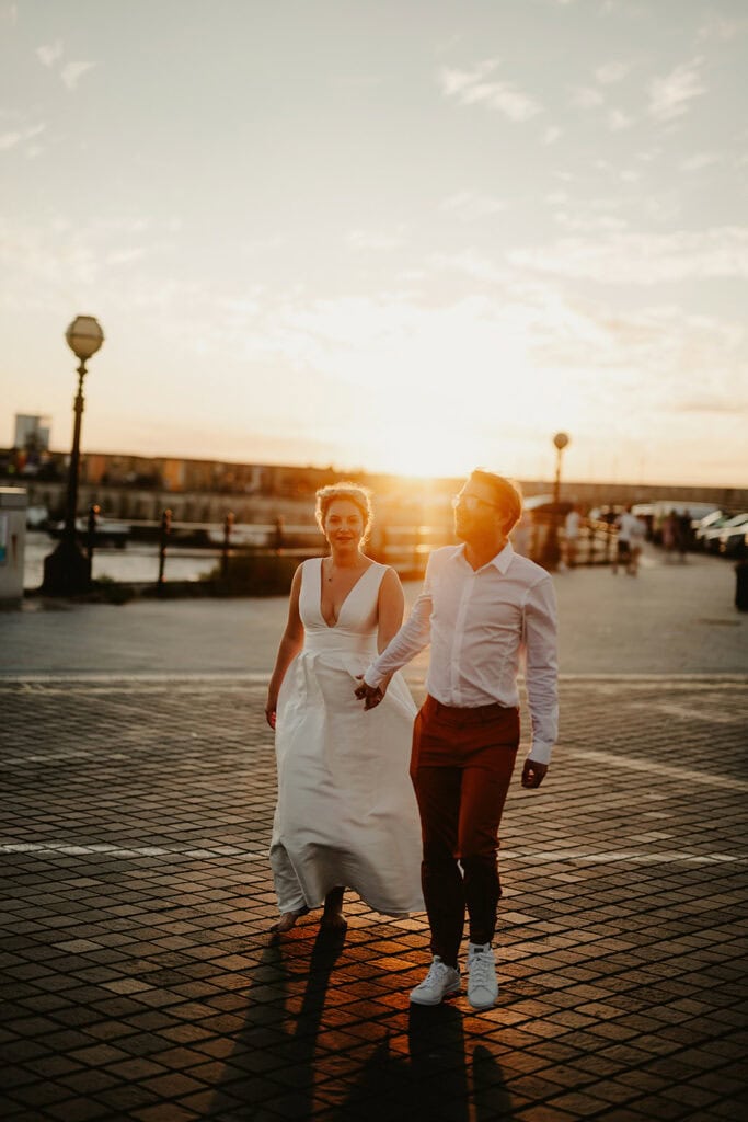 A couple walks hand-in-hand on a paved waterfront promenade during sunset. The woman wears a white dress, and the man wears a white shirt and red pants.